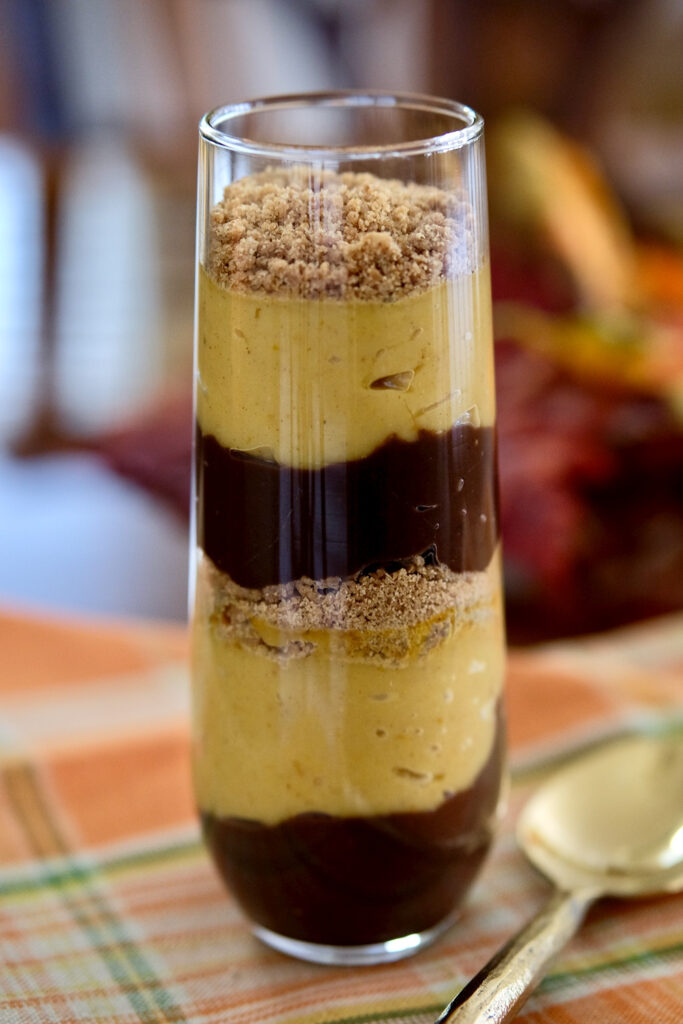 Chocolate pudding layered with pumpkin pudding and edible sand in a tall parfait glass set on a orange plaid napkin with gold spoon in forefront.  Walnut table and autumnal leaves are in background>