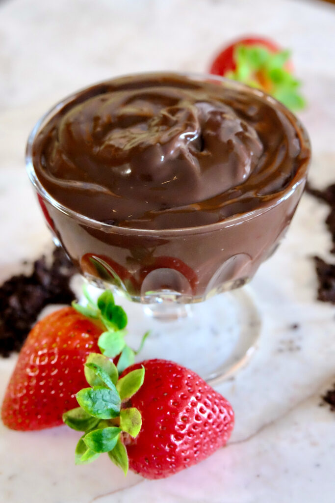Chocolate pudding in a stemmed glass dessert bowl set on white marble.  Chocolate sand is scattered around base with whole fresh strawberries. 