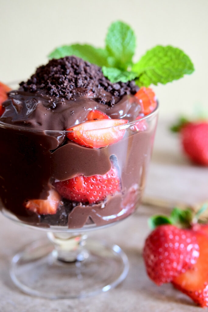 Glass dessert dish is layered with chocolate pudding, fresh strawberries and chocolate sand set on beiger marble top.   The dessert is garnished with additional chocolate sand and fresh mint. Fresh strawberries and spoon are in background.