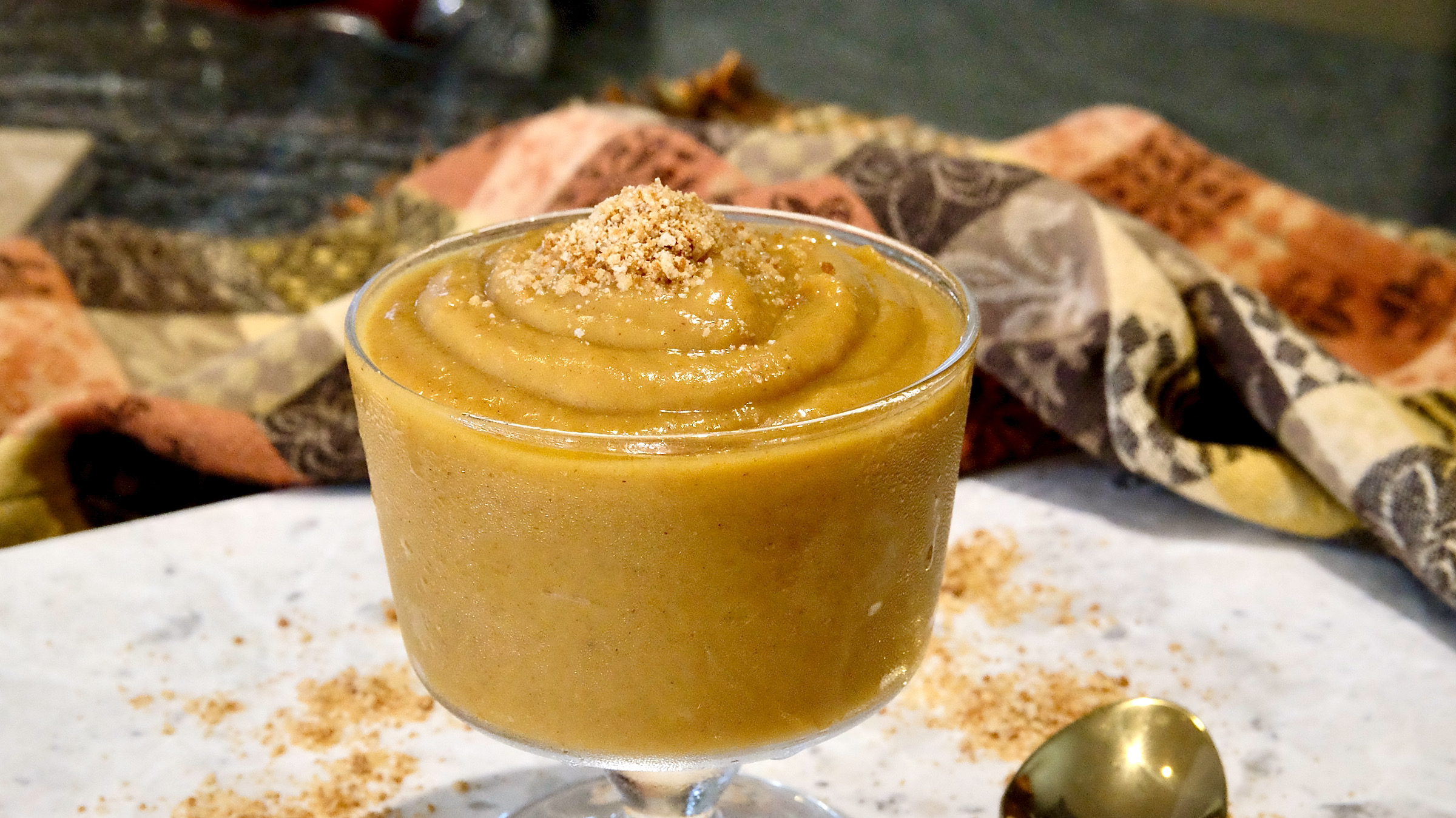 Pumpkin Pudding in a glass trifle bowl garnished with edible sand is set on white marble counter. Cookie sand is scattered around base of dessert with a gold spoon in forefront. Brown, beige and orange damask linen is in background.