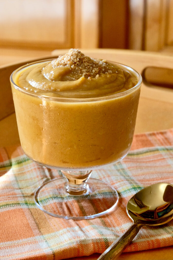 Pumpkin pudding in a glass dessert dish garnished with edible sand is set on a orange and green linen draped over a maple serving tray.  A  gold spoon is in right fore front.