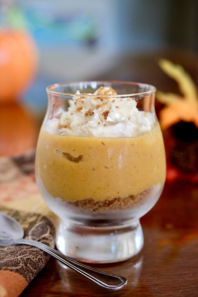 Edible sand and pumpkin pudding layered in a glass dessert cup is garnished with whipped cream sprinkled with edible sand.  Dessert cup is sitting on walnut wood table with autumn colored napkin in background with spoon.