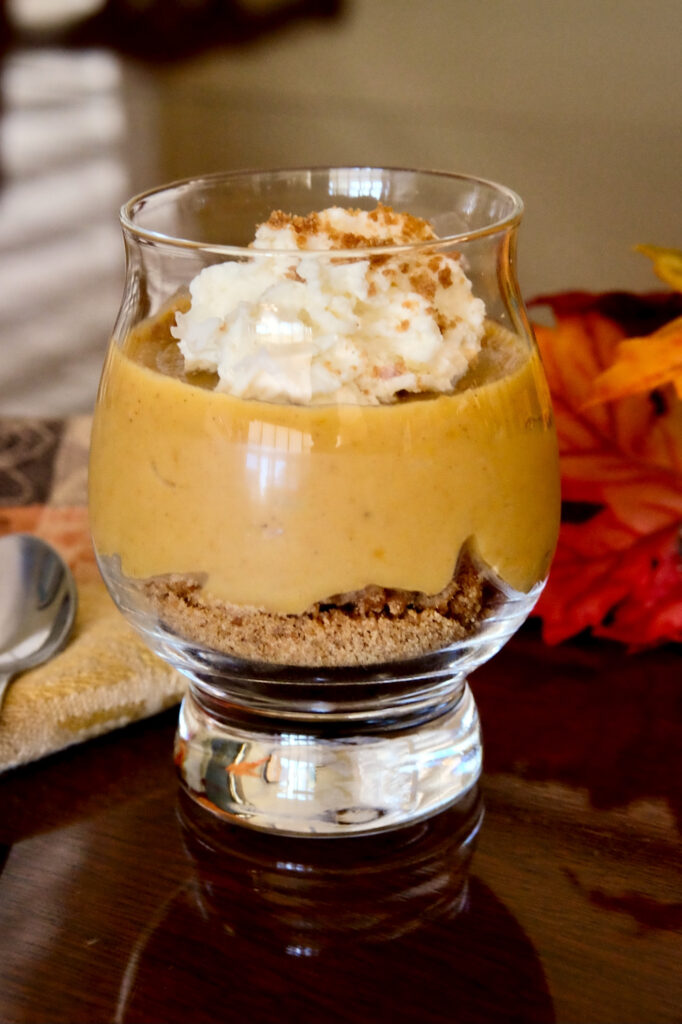 Pumpkin pudding layered on top of an edible cookie base in a glass dessert bowl.  Dessert is garnished with a dollop of whipped cream garnished with a sprinkling of edible sand.  Spoon is set on a Brown and tan linen in left background with red and orange autumn leaves in right side background>