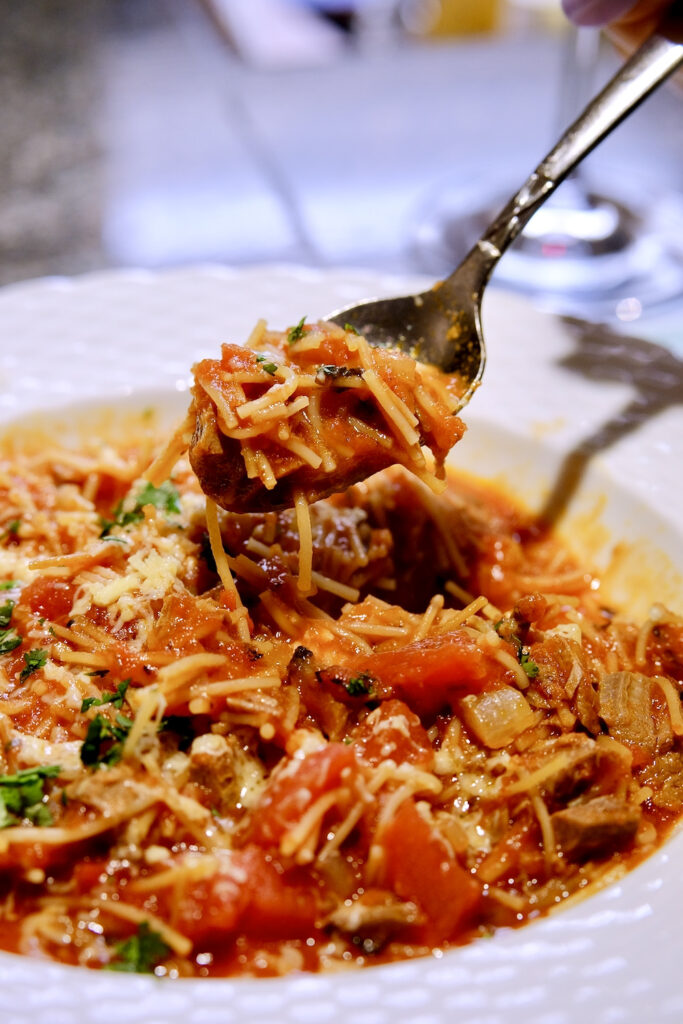 Thick tomato soup with chicken, noodles and vegetables served in a white rimmed bowl.  Spoon hovering over bowl showing a scoop of this hearty chunky dish.  