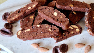 Double Chocolate biscotti scattered across white marble surface. Chocolate feves and almonds are scattered through fore and back ground of shot on marble surface.