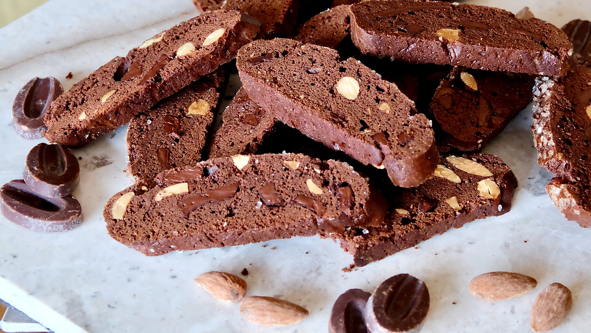 Double Chocolate biscotti scattered across white marble surface. Chocolate feves and almonds are scattered through fore and back ground of shot on marble surface.