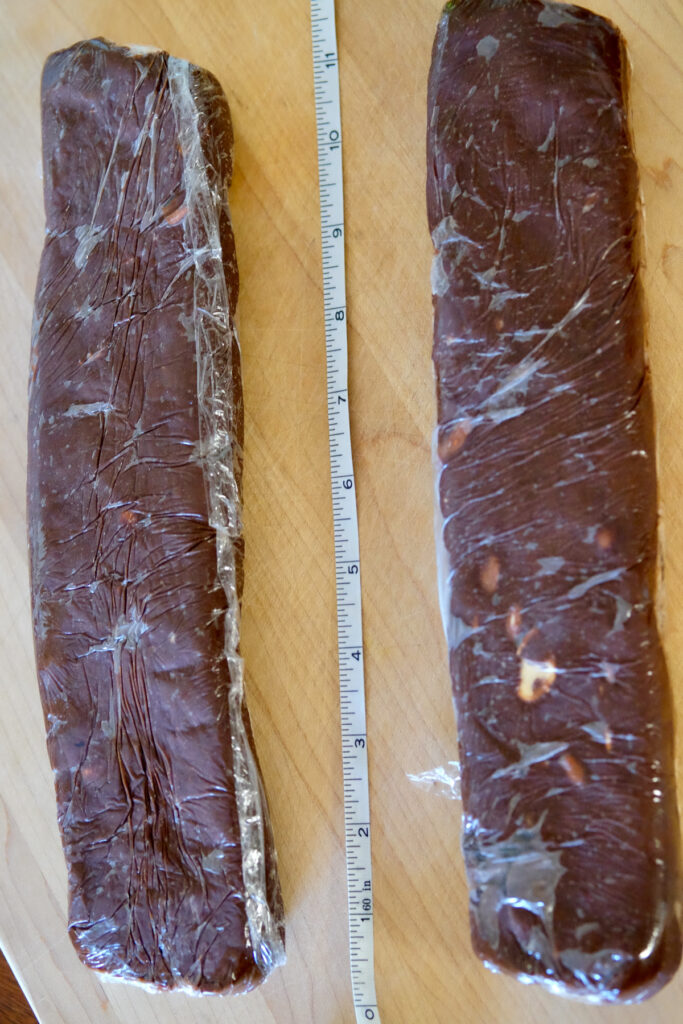 Chocolate biscotti wrapped and formed into log set on a maple wood board. Measuring tape between two logs depicting length of logs.
