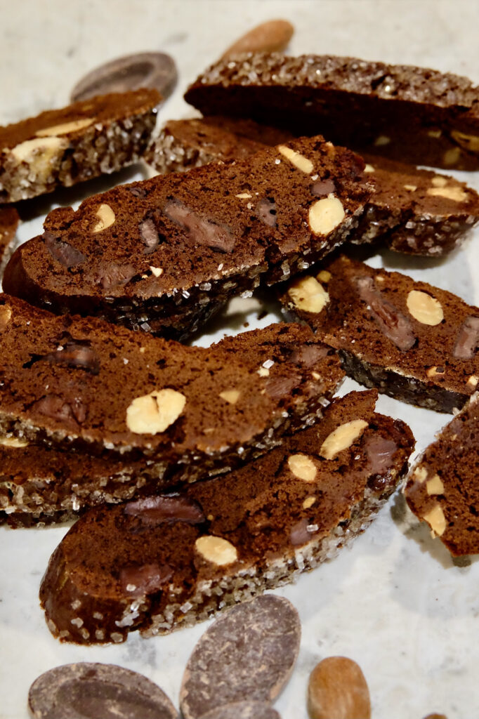 Double chocolate biscotti scattered on a white marble surface with chocolate feves and almonds scattered in fore and background.