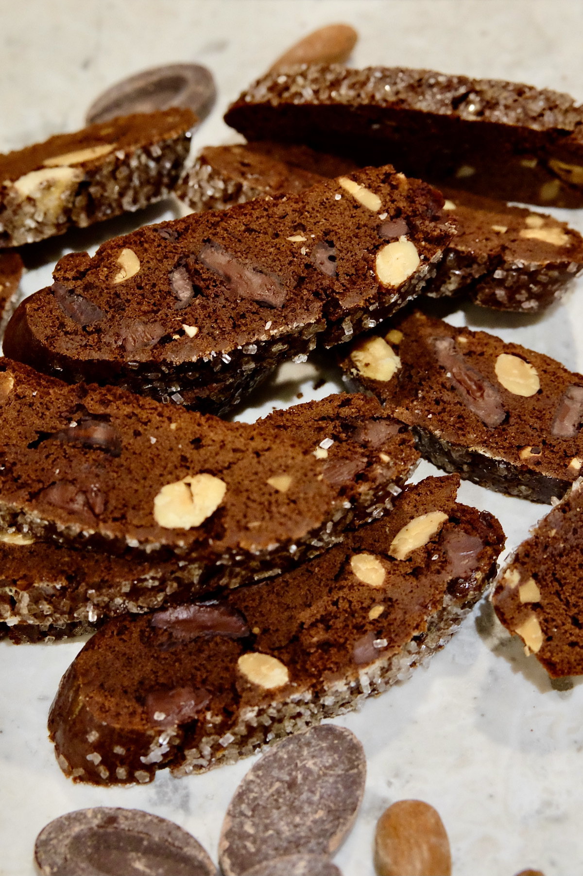 Chocolate Biscotti with Chocolate Ribbons and Almonds scattered on white marble counter with Chocolate Feves and Almonds scattered in fore and background.