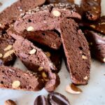 Chocolate Biscotti with dark chocolate ribbons and almonds are stacked on a white marble surface with chocolate disks and almonds scattered in foreground.