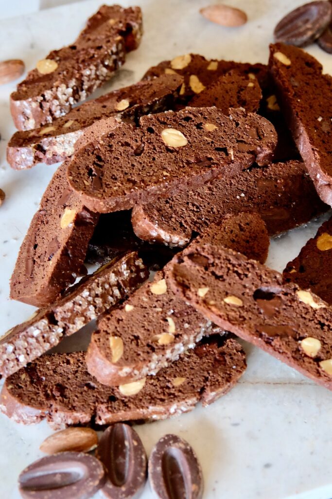 Double Chocolate Biscotti stacked on white marble counter with chocolate pieces and almonds in foreground.