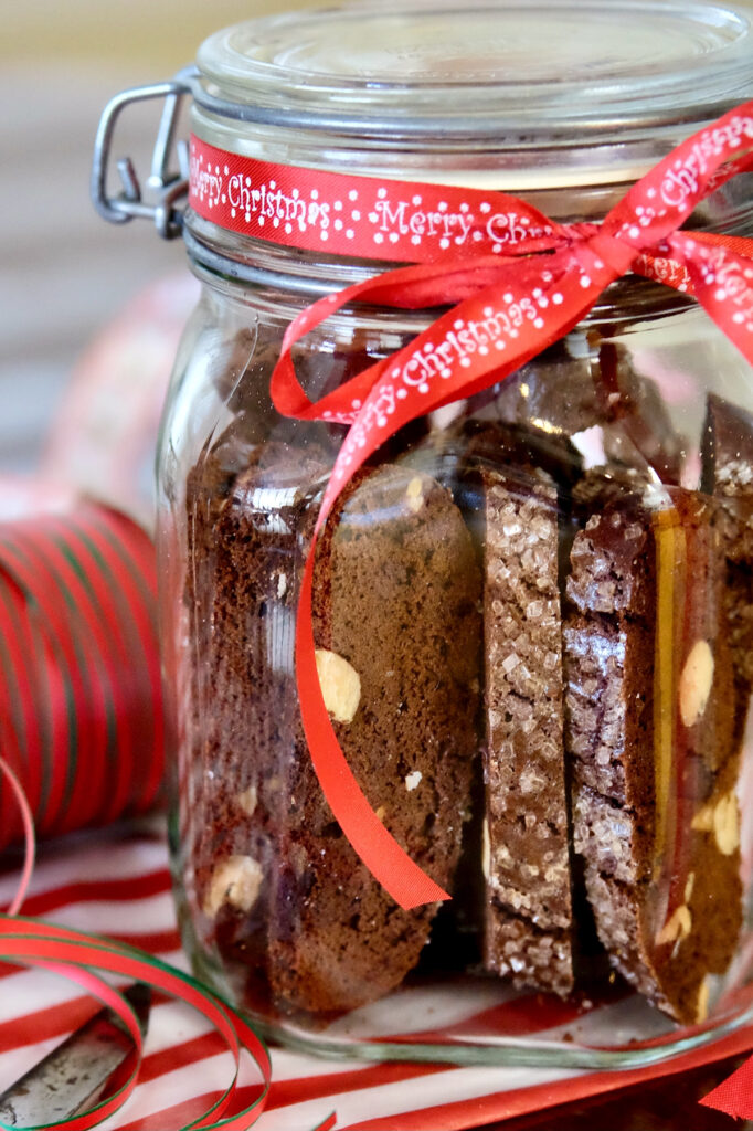 Double Chocolate Biscotti stored in a glass jar with red ribbon tied around jar is set on red and white striped paper with scissors and spool of ribbon in foreground.