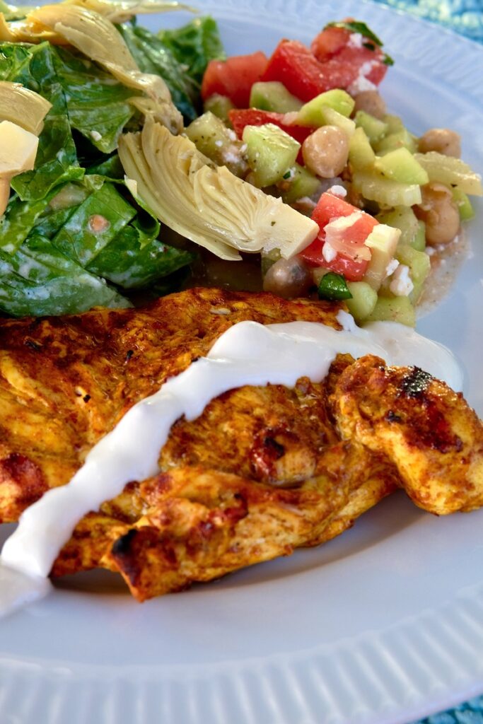 Chicken Shawarma drizzled with yogurt sauce served on a white dinner plate with Tomato Cucumber Salad and Green Mediterranean Salad with Creamy Greek Dressing