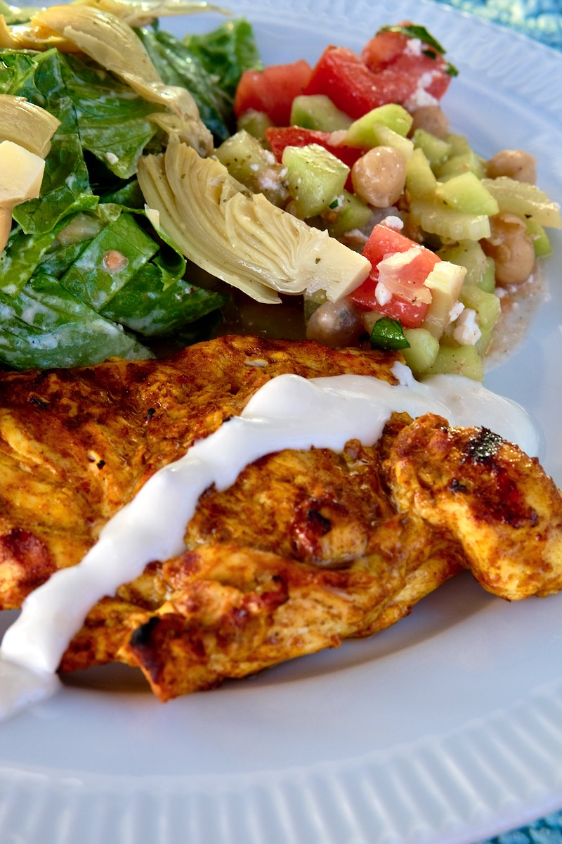 Chicken Shawarma with Yogurt Sauce served on forefront of white plate with Tomato Cucumber Salad in upper right  and Mediterranean  Green Salad in upper left corner of plate.