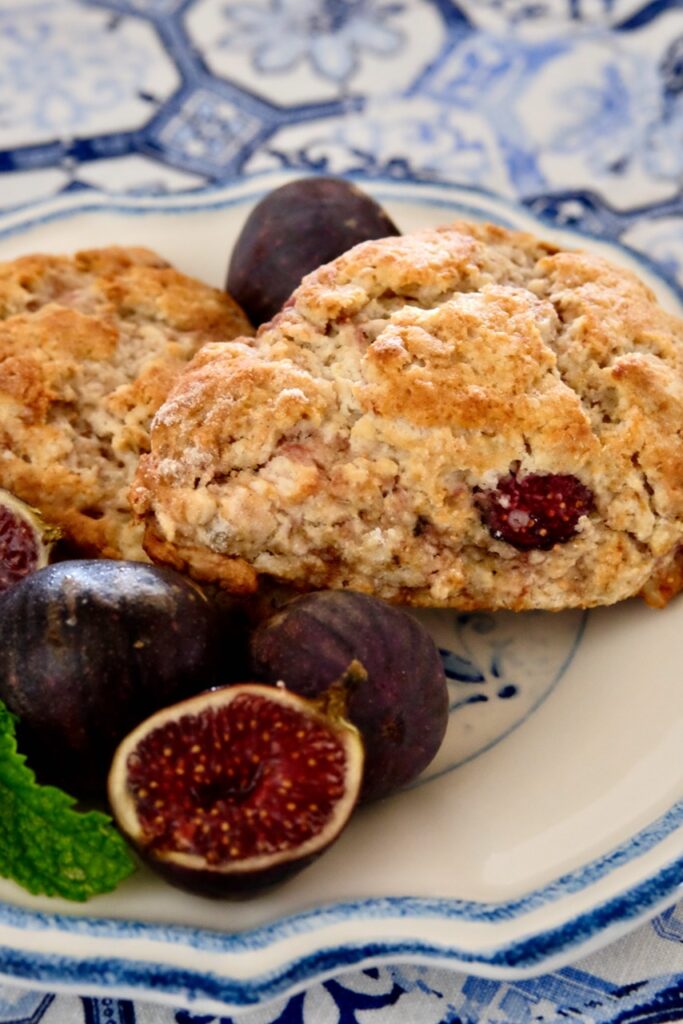 Two fig scones served on a white and blue rimmed plate set on white and blue tiled linen.  Fresh whole figs and cut figs are displayed in fore front of plate.