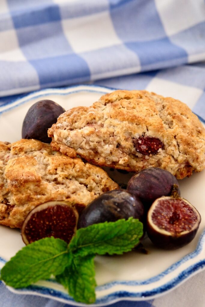 Two fresh fig scones served on blue and white rimmed plate set on blue and white plaid linen.  Whole and cut figs are displayed on the forefront of plate with mint sprig.