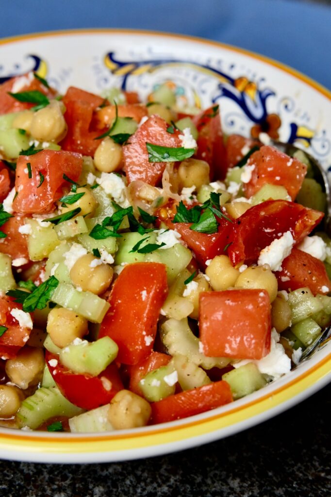 Tomatoes cucumber salad in a multi-colored Italian serving bowl is tossed with garbanzo beans and crumbled feta cheese and garnished with chopped parsley.  Bowl is set on a blue linen cloth. 