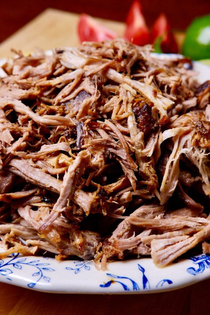 Shredded pork carnitas in a white and blue serving bowl with tomato wedges and lime in background.