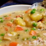 A multi colored soup bowl filled to the brim with chicken pot pie soup while a spoonful hovers over the bowl.