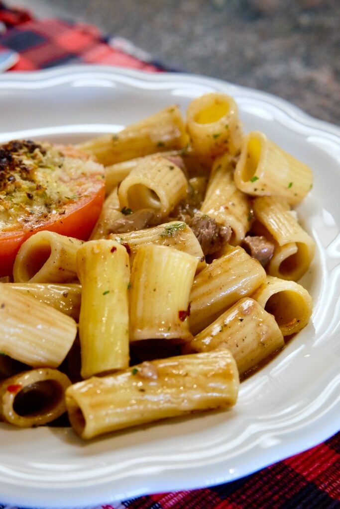 Rigatoni pasta  baked with cubes of roasted meat, gravy and herbs served in a white ruffle edged bowl.  A stuffed tomato is peaking out of the pasta in upper left corner of bowl.