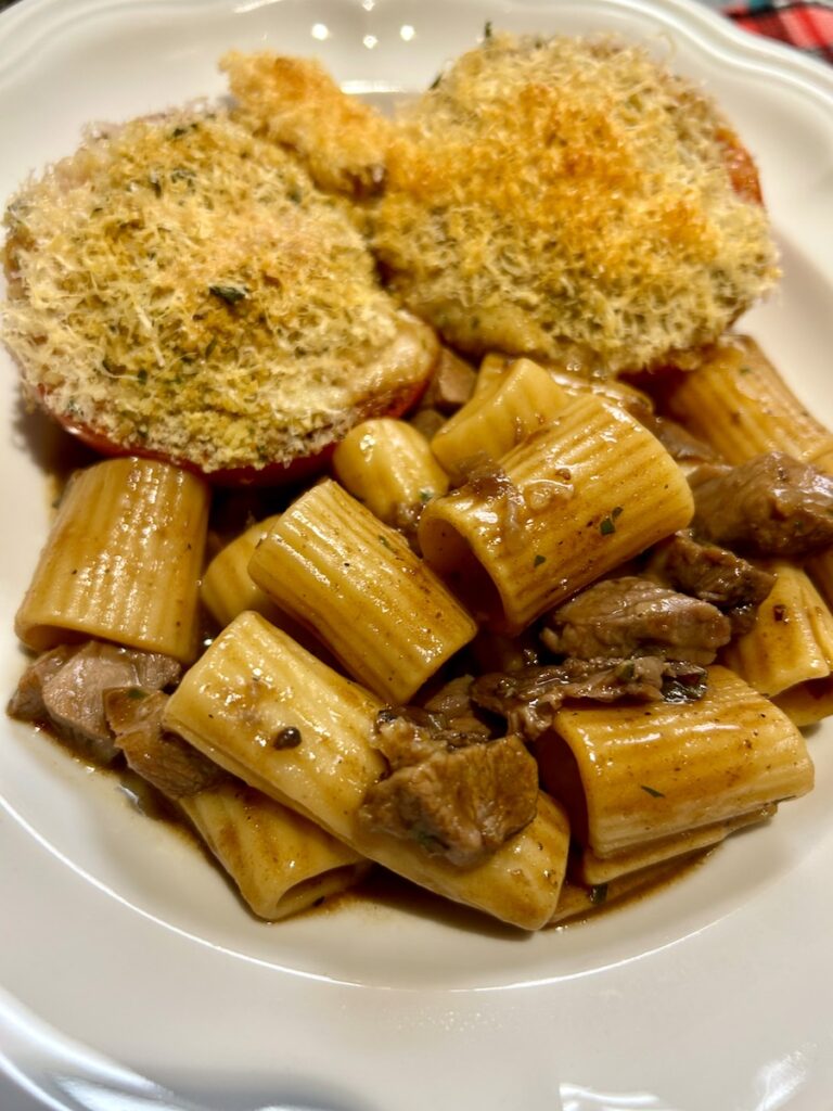 Rigatoni pasta baked with gravy and leftover cubed roast, served in a white pasta bowl with two stuffed tomatoes at the top of the bowl.